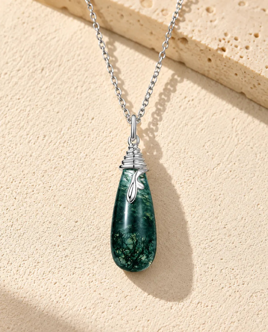 Moss Agate Halsband med Droppe i 925 Sterlingsilver - liggande på en ljus strukturerad yta bredvid ett travertinblock