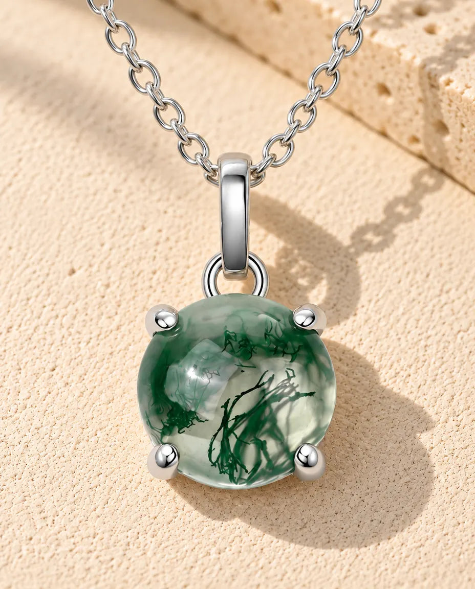 Moss Agate Halsband med Rund Sten i 925 Sterlingsilver - liggande på en ljus strukturerad yta bredvid ett travertinblock