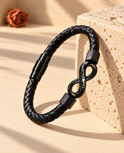 Svart armband i flätat konstläder med graverad oändlighetssymbol, lutat mot ett block av travertin