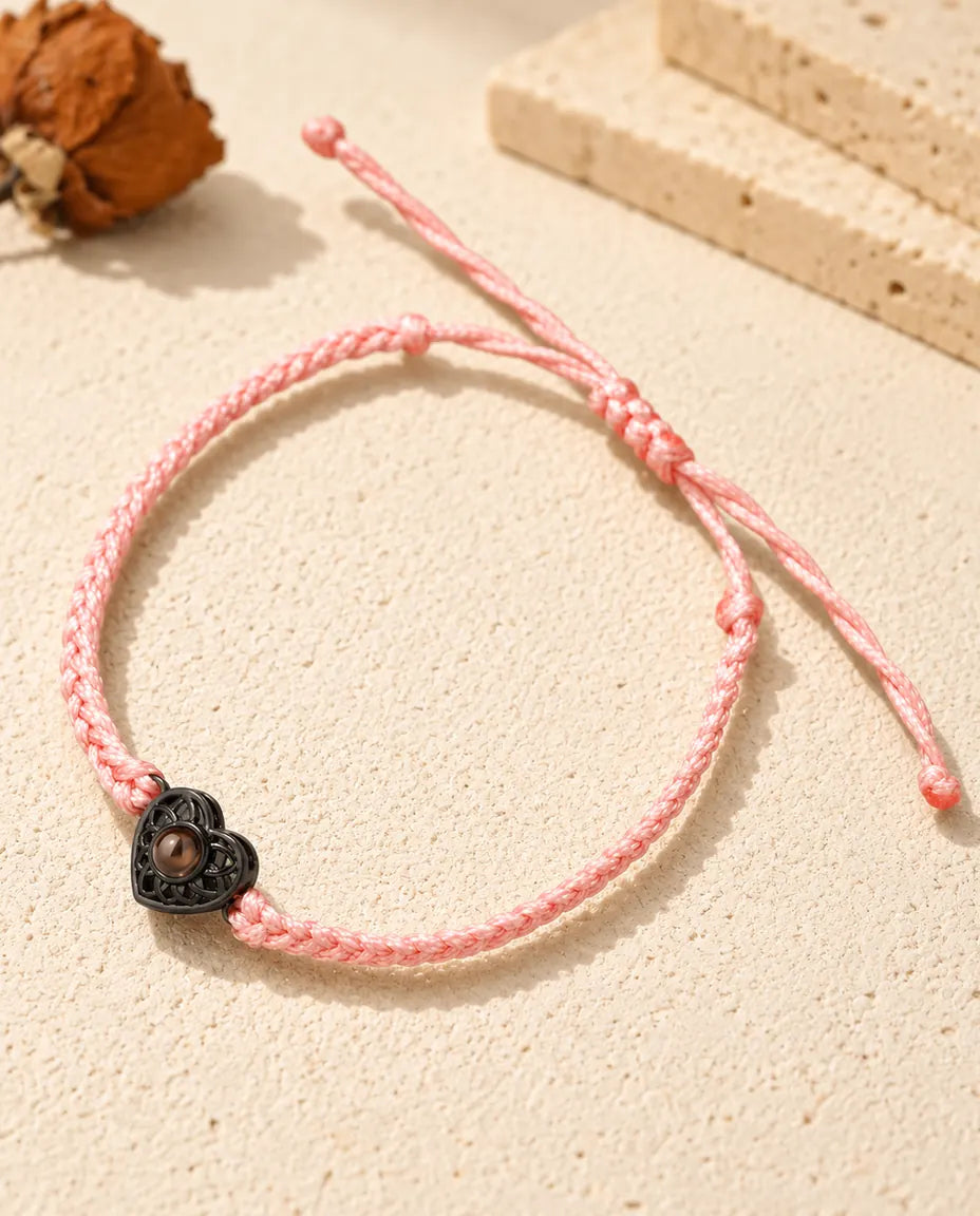 Armband med rosa tunn flätad snodd och svart hjärtberlock som håller en brun projektionssten