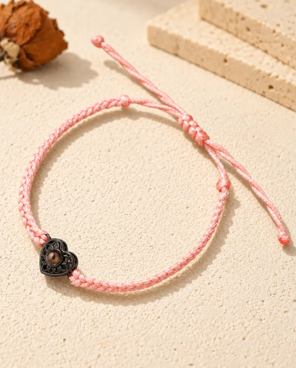 Armband med rosa tunn flätad snodd och svart hjärtberlock som håller en brun projektionssten