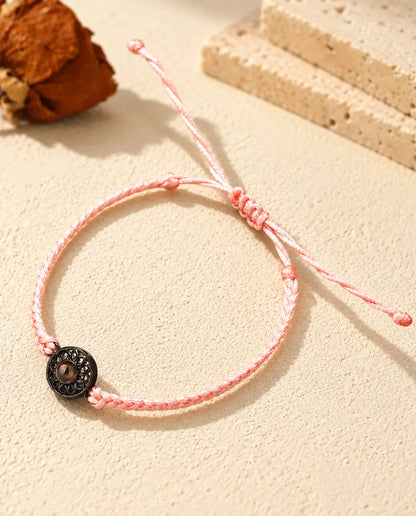 Armband med rosa tunn flätad snodd och svart rund berlock som håller en brun projektionssten
