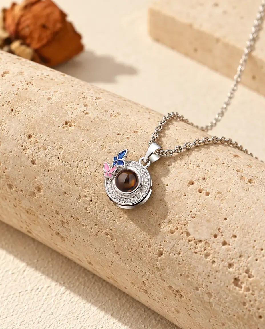 Halsband med bild inuti i silverfärgat utförande, runt hänge med mörkblå och rosa emaljfjärilar, på travertin