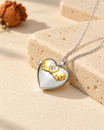 Personlig silverfärgad hjärtmedaljong med guldfärgade änglavingar på locket, lutad mot ett travertinblock