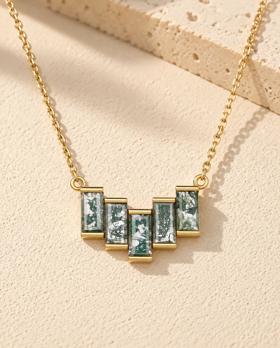 Moss Agate Halsband i Guldpläterat 925 Sterlingsilver - liggande på en ljus strukturerad yta bredvid ett travertinblock