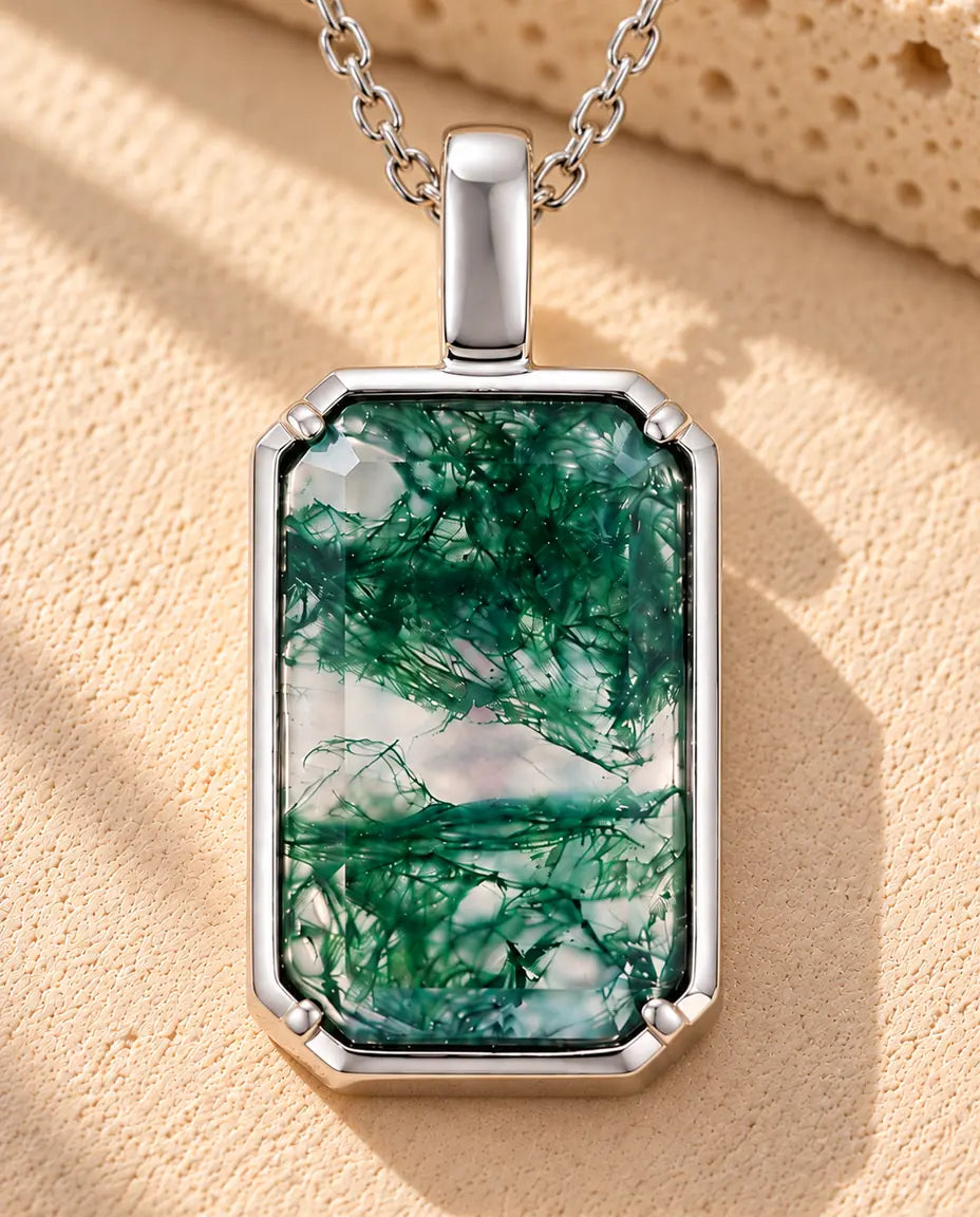 Moss Agate Halsband med Rektangulärt Hänge i 925 Sterlingsilver - liggande på en ljus strukturerad yta bredvid ett travertinblock