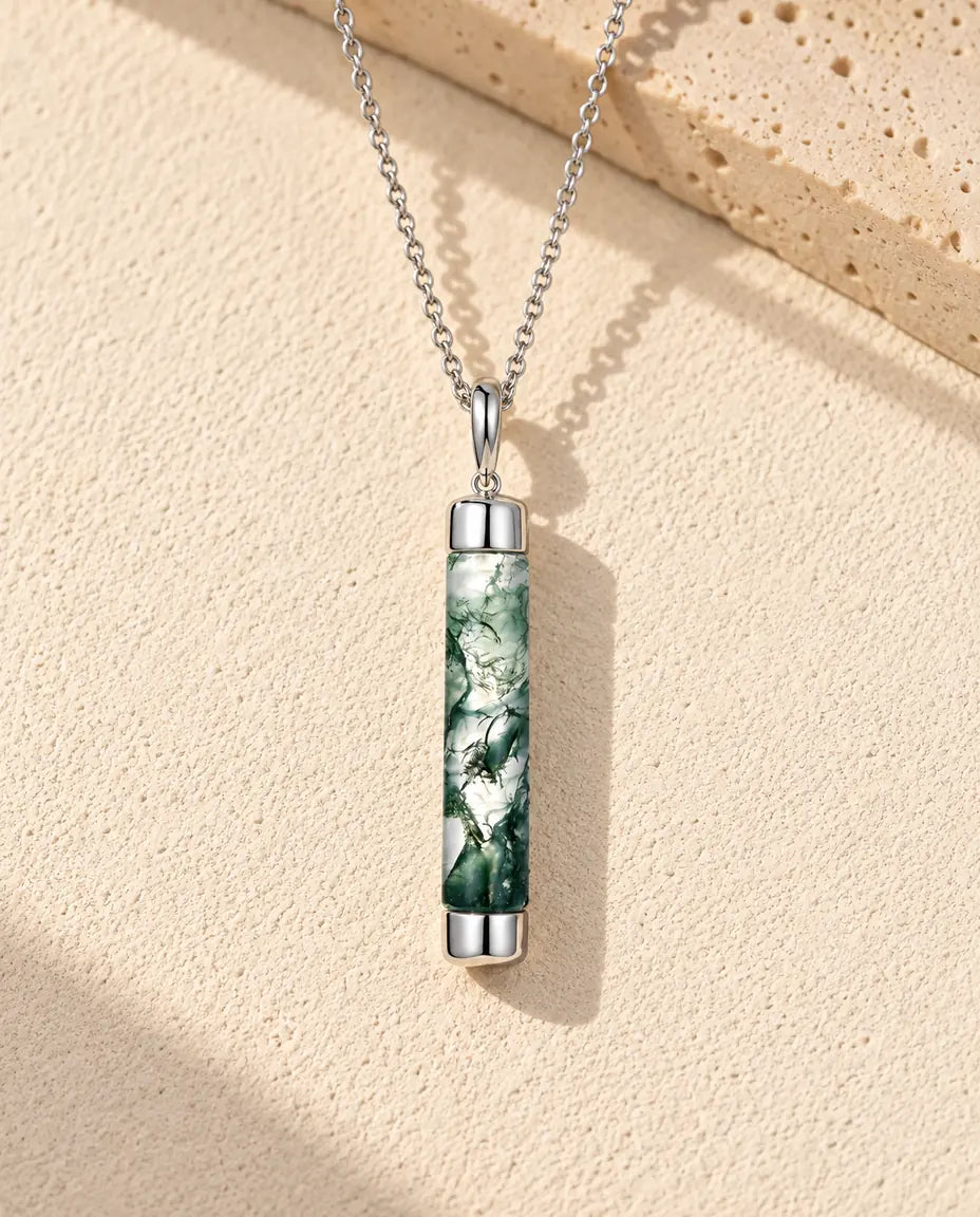 Moss Agate Halsband med Lång Cylinder i 925 Sterlingsilver - liggande på en ljus strukturerad yta bredvid ett travertinblock