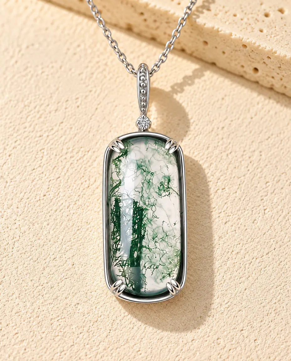 Moss Agate Halsband med Rektangulärt Hänge i 925 Sterlingsilver - liggande på en ljus strukturerad yta bredvid ett travertinblock