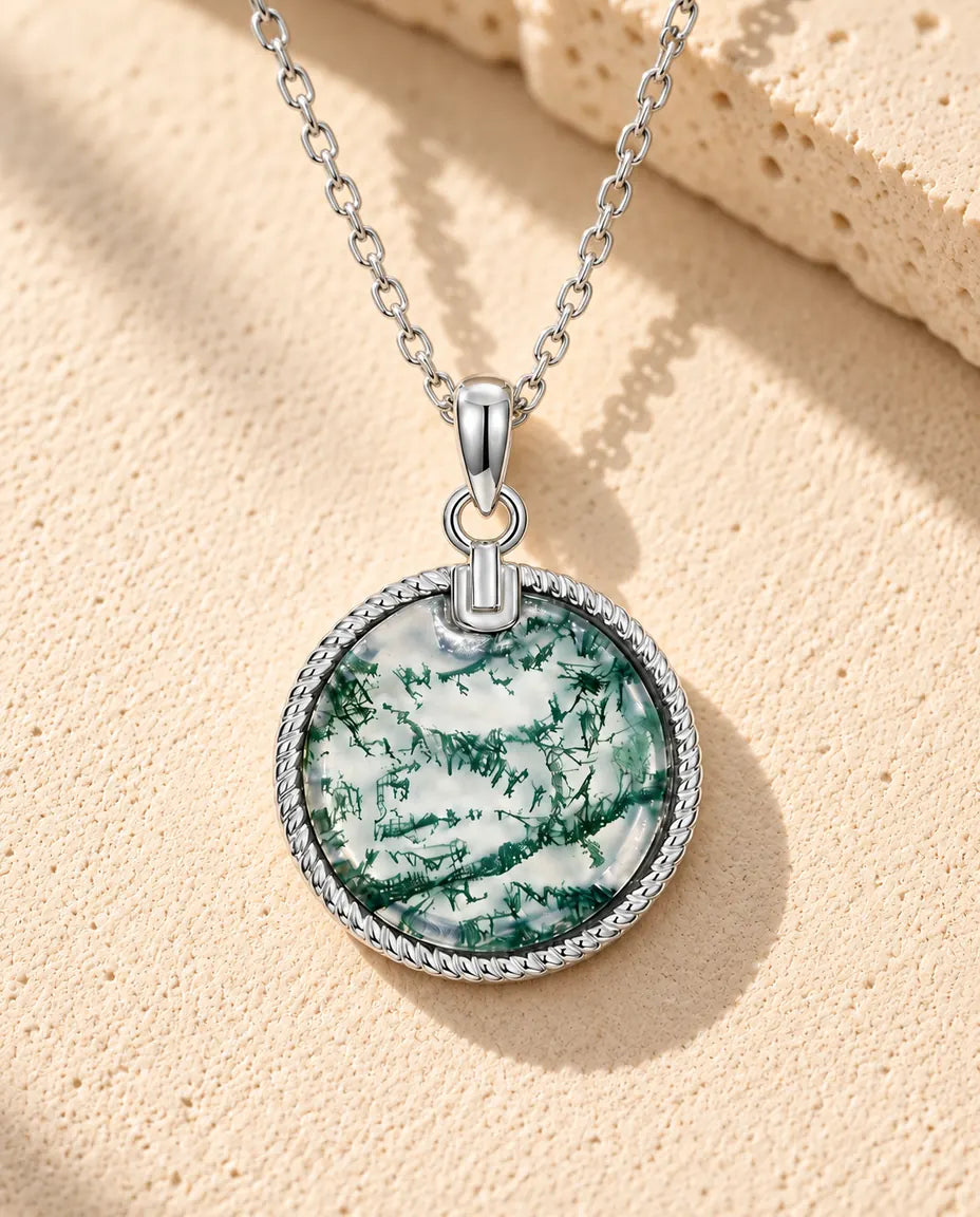 Moss Agate Halsband med Rund Medaljong i 925 Sterlingsilver - liggande på en ljus strukturerad yta bredvid ett travertinblock