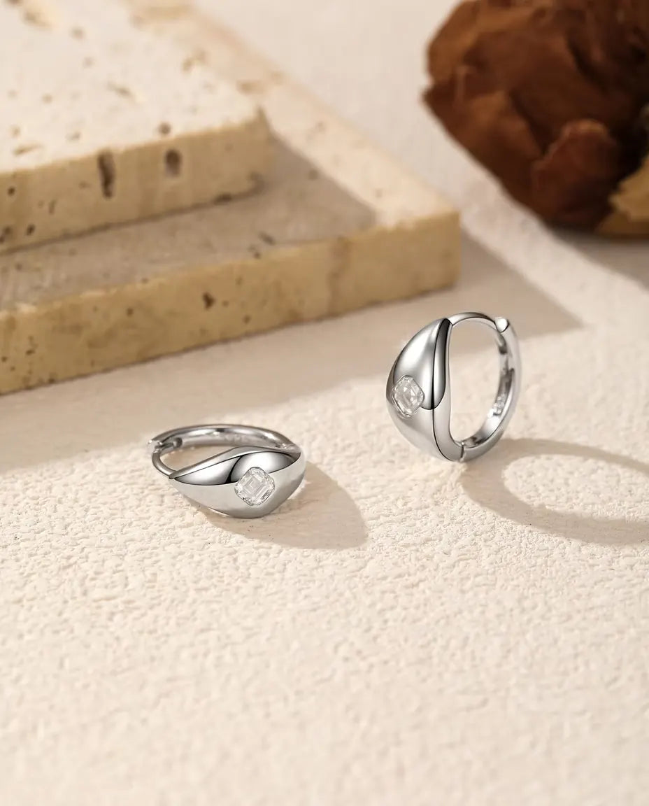 Moissanite Örhängen i 925 Sterlingsilver - paret på en ljus strukturerad yta, ett liggande och ett stående